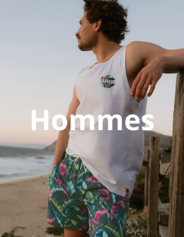 Homme près de la plage se tenant contre une clôture avec une planche de surf dans les mains, portant une camisole et un maillot de bain de la marque Hamabe.